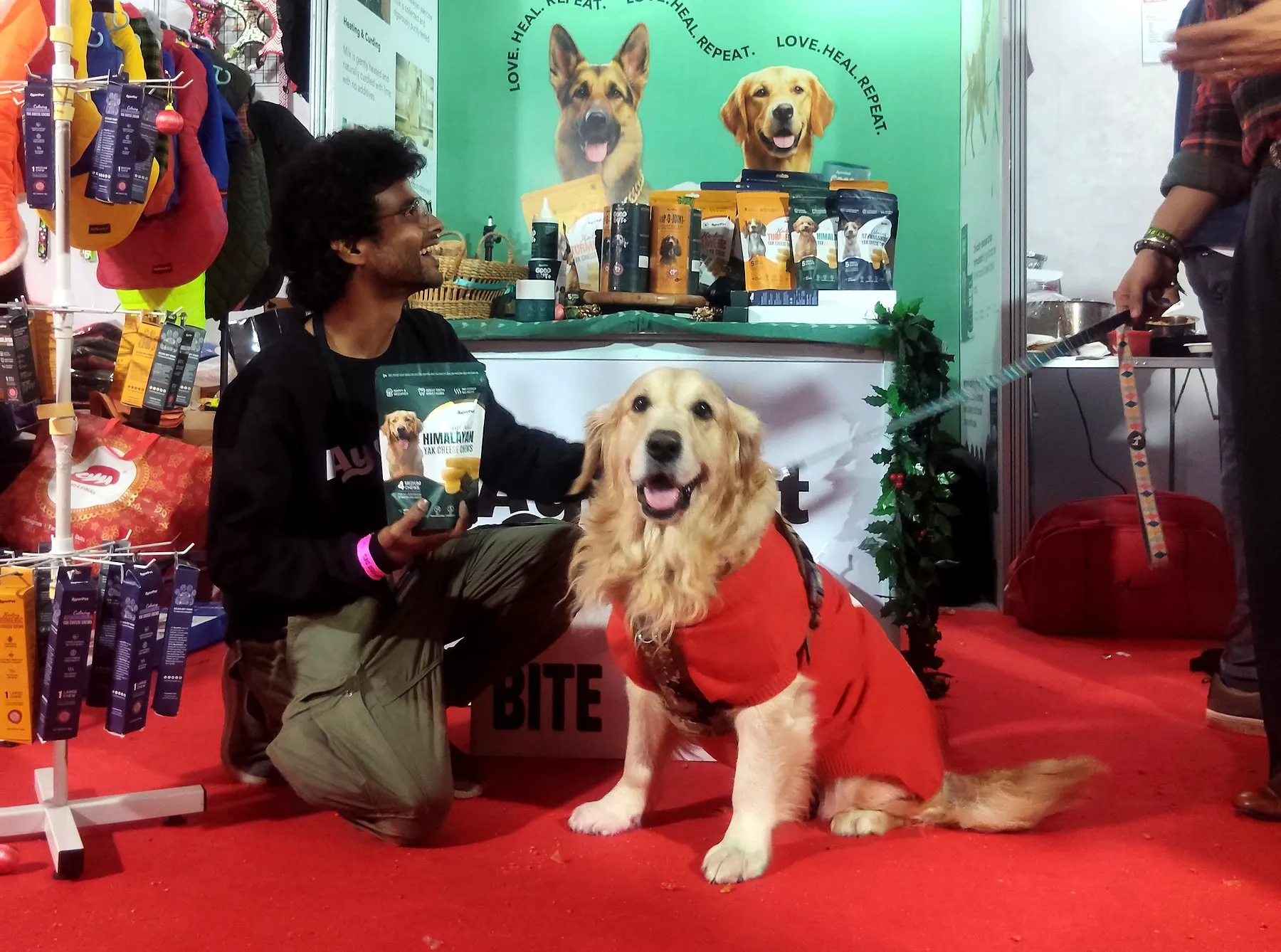 A team member kneeling beside a golden retriever in a red jacket at a pet expo stand